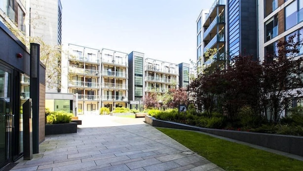 Ein moderner Innenhof in der Stadt mit einem gepflasterten Weg, umgeben von mehrstöckigen Wohngebäuden mit Balkonen und großen Fenstern.