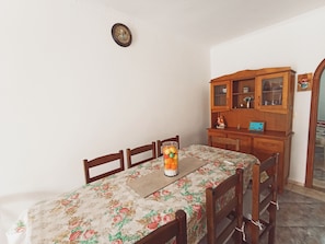 Uma sala de jantar com toalha de mesa florida, cadeiras de madeira, um armário com portas de vidro e um relógio de parede.