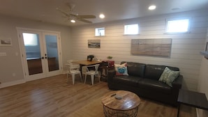 A living room with a wooden floor, a black leather sofa, a round coffee table, a dining table with chairs, and a ceiling fan.