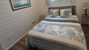 A bedroom with a bed, a window with curtains, a framed picture on the wall, and a small table with a lamp.
