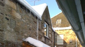 A stone house with a snow-covered roof and a wooden porch. - Quaint cottage in the heart of the village in the heart of Scotland. (Aberfeldy)