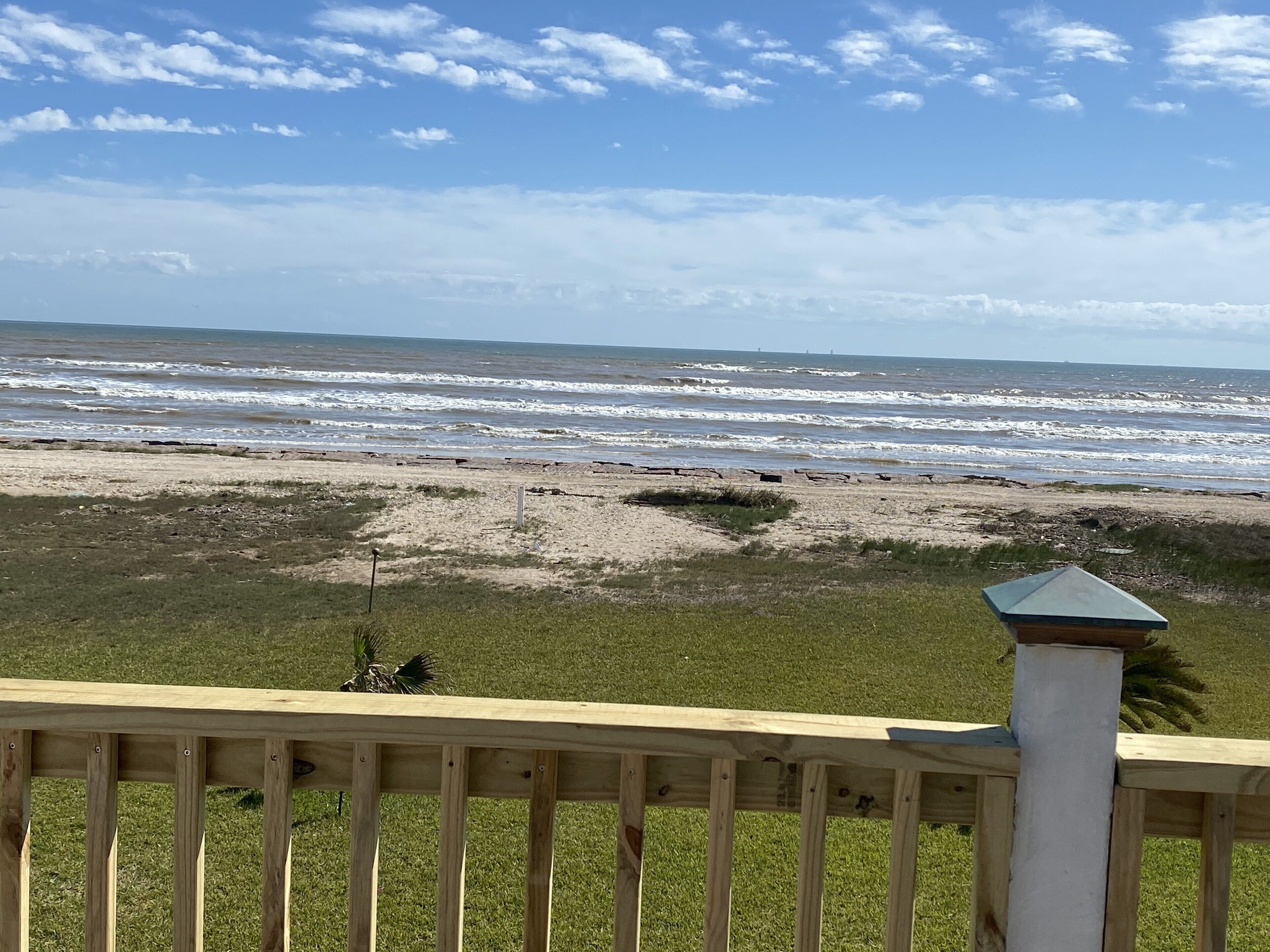 Vue sur la plage depuis une terrasse en bois dotée d’une balustrade, avec une zone herbeuse et les vagues de l’océan en arrière-plan.