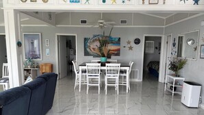 Un vaste espace de vie comprenant une table à manger, des chaises blanches, un ventilateur de plafond et un grand tableau.