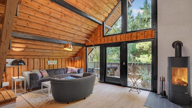 Un interior de cabaña acogedor con techo de madera, un sofá gris, una mesita y una chimenea.