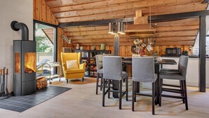 Interior acogedor de una cabaña con estufa de leña, comedor con sillas, cocina con ollas colgantes y una estantería.