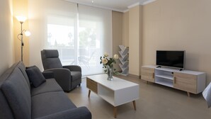 A modern living room with a grey sofa, a white coffee table with a vase of flowers, a flat-screen TV on a white TV stand, and a large window with a view of palm trees. - Brisas B2, 2 bedrooms, beach at 50m, by Bookindenia (Dénia)