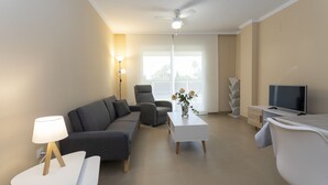 A modern living room with a grey sofa, a white coffee table, a TV, and a ceiling fan. - Brisas B2, 2 bedrooms, beach at 50m, by Bookindenia (Dénia)