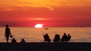Una puesta de sol sobre una playa con gente sentada en sillas y caminando por la orilla.