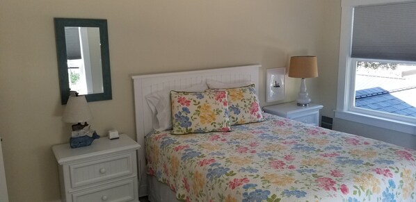 A bedroom with a bed, a nightstand, a lamp, a mirror, and a window with a view of the outside.