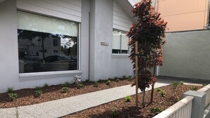 Un bâtiment moderne à l’extérieur blanc, doté d’un chemin pavé et d’un petit espace jardin agrémenté de paillis et de jeunes plantes.