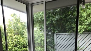 Une vue à travers une grande fenêtre donnant sur une terrasse en bois, un pot de fleurs et une clôture blanche.