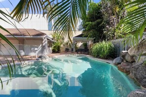 Una piscina circondata da rocce e piante, con una zona patio coperta e un edificio sullo sfondo.