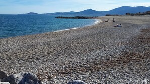 Una playa de guijarros con un rompeolas rocoso, montañas a lo lejos y cielos despejados.