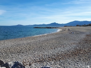 A pebbled beach with a rocky breakwater, distant mountains, and clear skies.