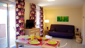 Un área de comedor con una mesa preparada para dos personas, un televisor sobre un mueble bajo y un sofá al fondo.