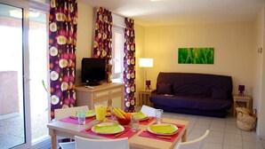 A dining area with a table set for two, a television on a stand, and a sofa in the background.