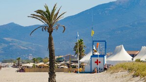 A beach with a palm tree, a lifeguard tower, and tents in the background.