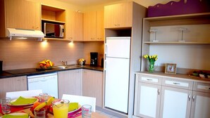 A modern kitchen with wooden cabinets, a microwave, a coffee maker, and a refrigerator. There is a dining table set with plates, glasses, and a fruit bowl. - Appartement confortable avec terrasse/loggia privée | accès Piscine (Saint-Cyprien)