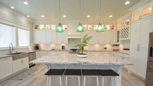 Een moderne keuken met witte kasten, een groot eiland met barstoelen, hanglampen en een plant in het midden.