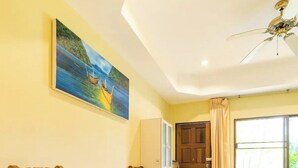 A room with a wooden dining table and chairs, a painting on the wall, a ceiling fan, a window with curtains, and a bed with a green cover. - Baan Coconut (Choeng Thale)