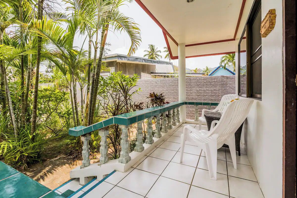 A covered balcony with white plastic chairs, a tiled floor, and a view of a building and greenery.