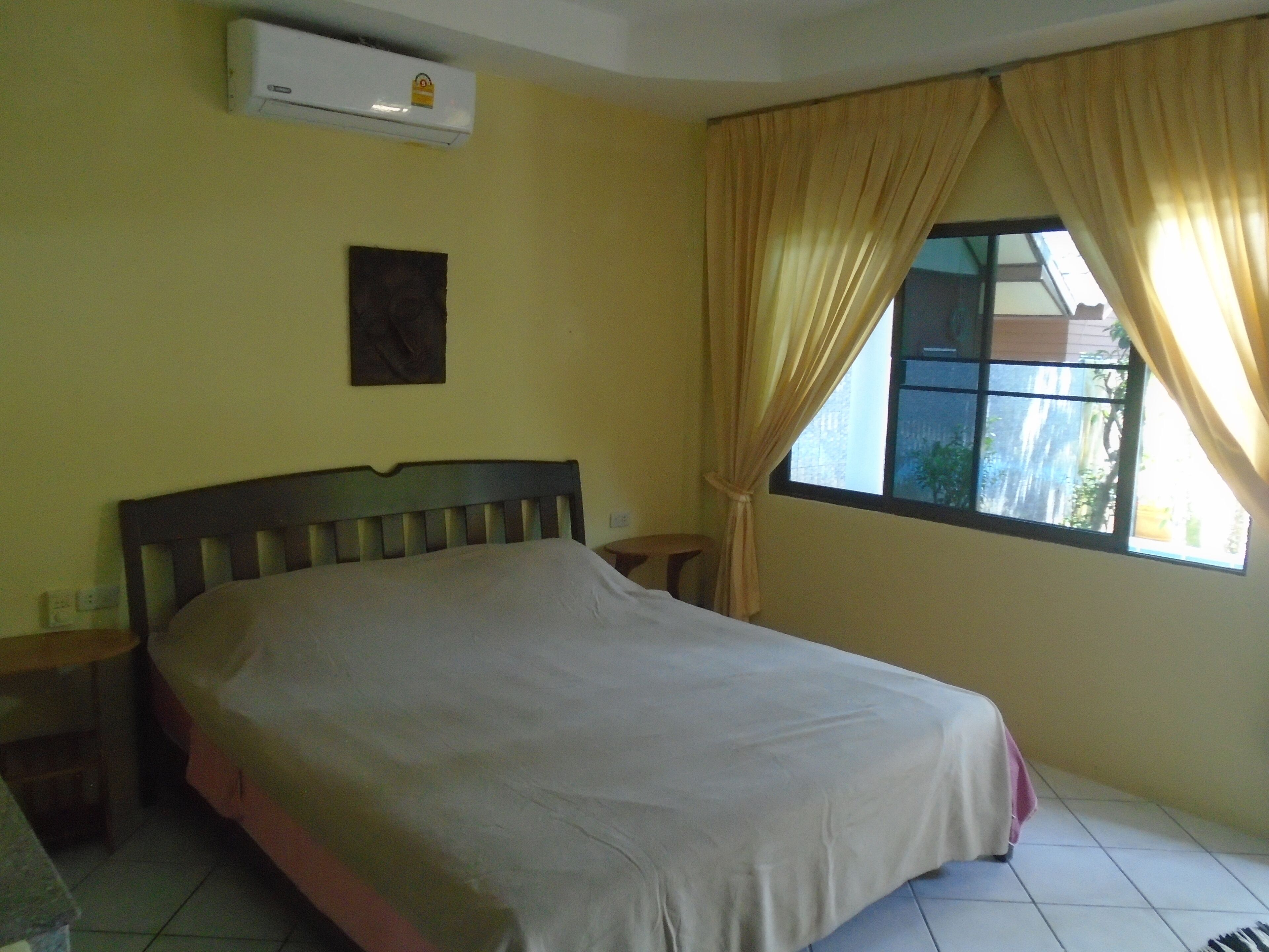 A bedroom with a bed, an air conditioner, a window with curtains, and a small table.
