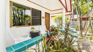 A covered outdoor seating area with white plastic chairs, a small round table with a vase of flowers, and a view of tropical vegetation through a window. - Baan Coconut (Choeng Thale)
