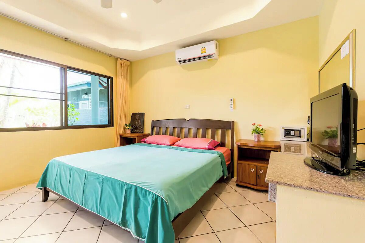 A hotel room with a bed, a television, a small table, a nightstand, an air conditioner, and a window with curtains.