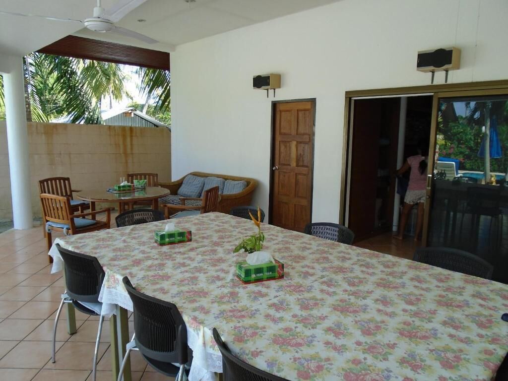 A dining area with a table covered in a floral tablecloth, surrounded by chairs. There is a wooden door and a sliding glass door leading to another room.