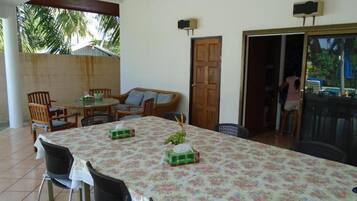A dining area with a table covered in a floral tablecloth, surrounded by chairs. There is a wooden door and a sliding glass door leading to another room.