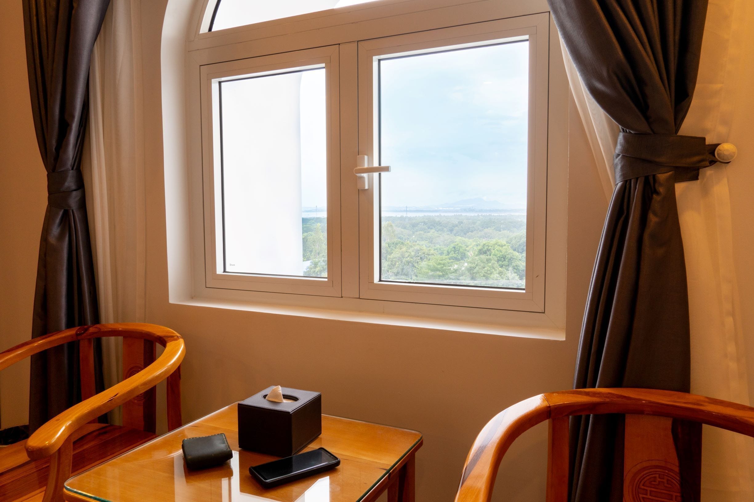 A room with a wooden table, a chair, a window with curtains, and a view of greenery outside.