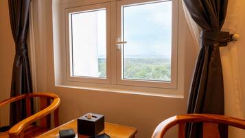 A room with a wooden table, a chair, a window with curtains, and a view of greenery outside.