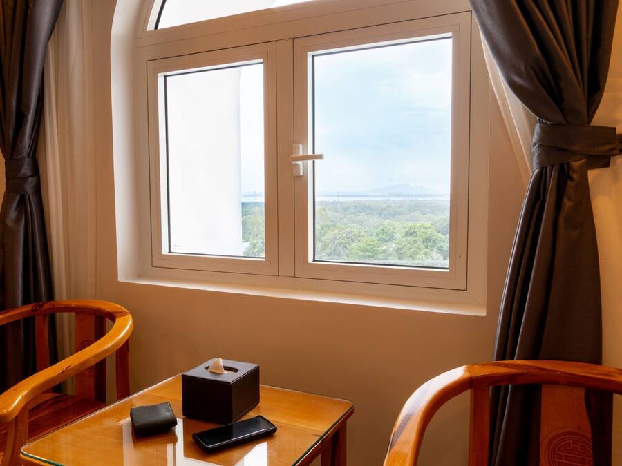 A room with a wooden table, a chair, a window with curtains, and a view of greenery outside.