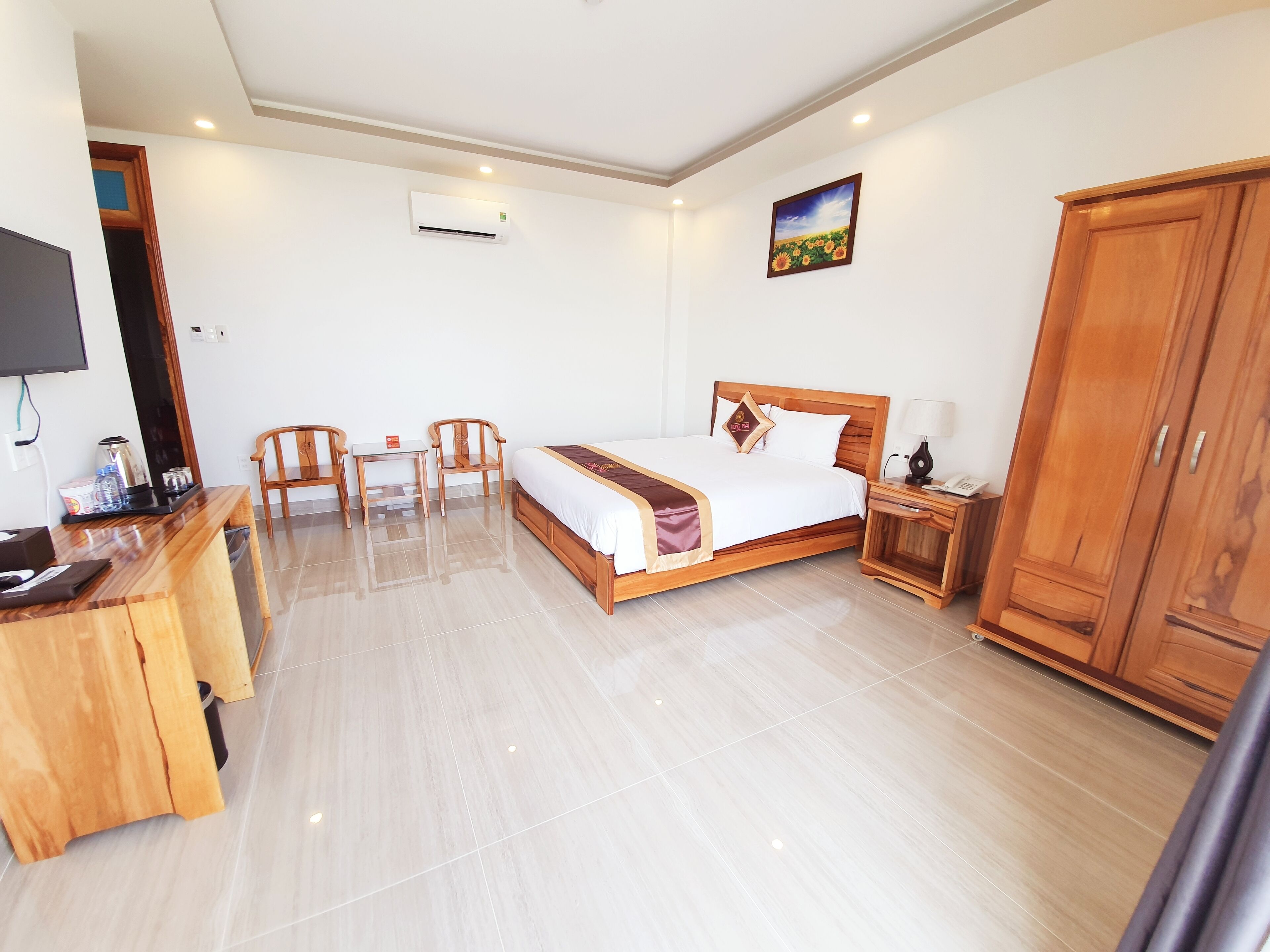 A hotel room with a bed, bedside table, lamp, wardrobe, desk with chairs, and a television.