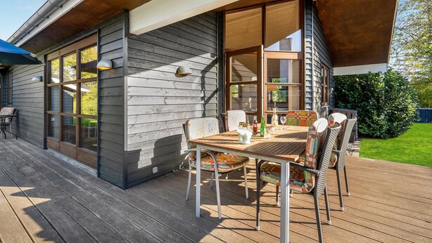 Eine Holzterrasse mit Tisch und Stühlen, großen Fenstern und Blick auf eine grüne Landschaft.