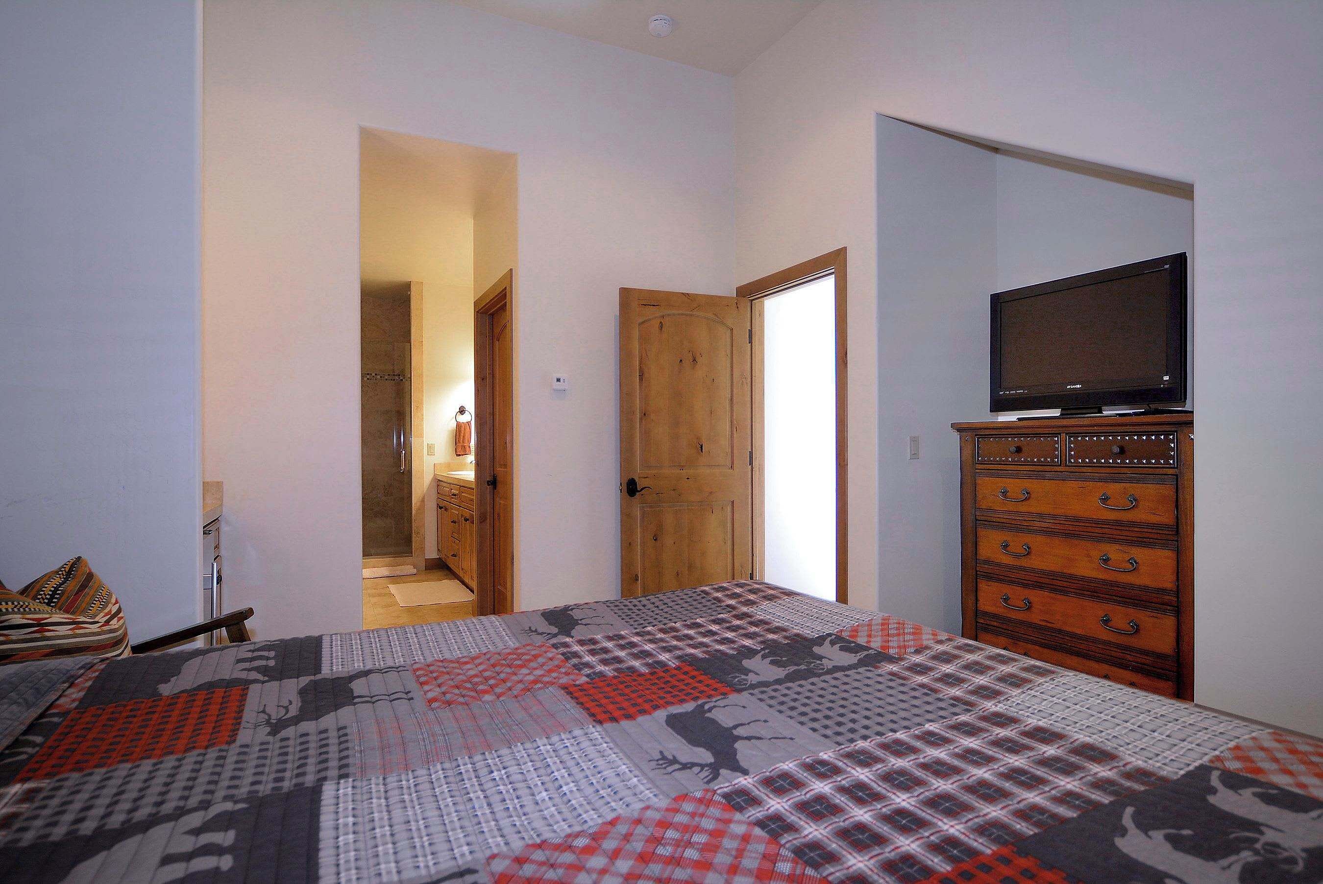 Un dormitorio con cama, tocador, televisión y baño visible a través de una puerta abierta.