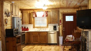 Interior de una cabaña de madera con cocina equipada: armarios de madera, refrigerador, microondas y lavavajillas. Cuenta con un comedor con mesa y sillas, y un televisor montado en la pared.