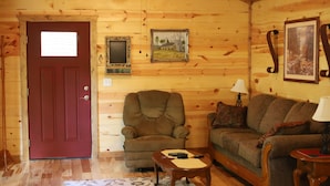 Interior acogedor de una cabaña de troncos con una puerta roja, un sillón marrón, un sofá y una mesita con un control remoto.