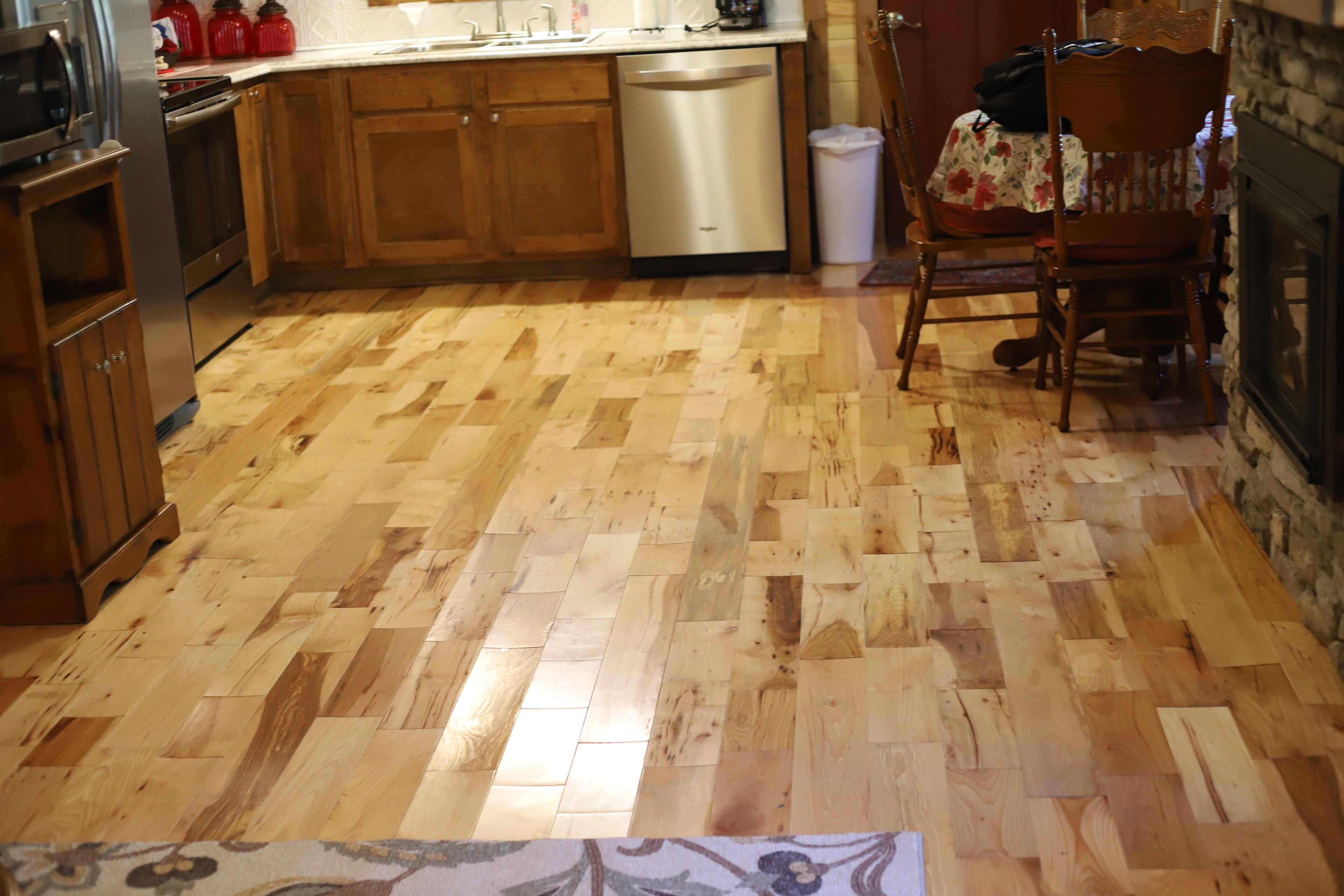 A kitchen with wooden flooring, wooden cabinets, a dishwasher, and a dining area with a chair and table.