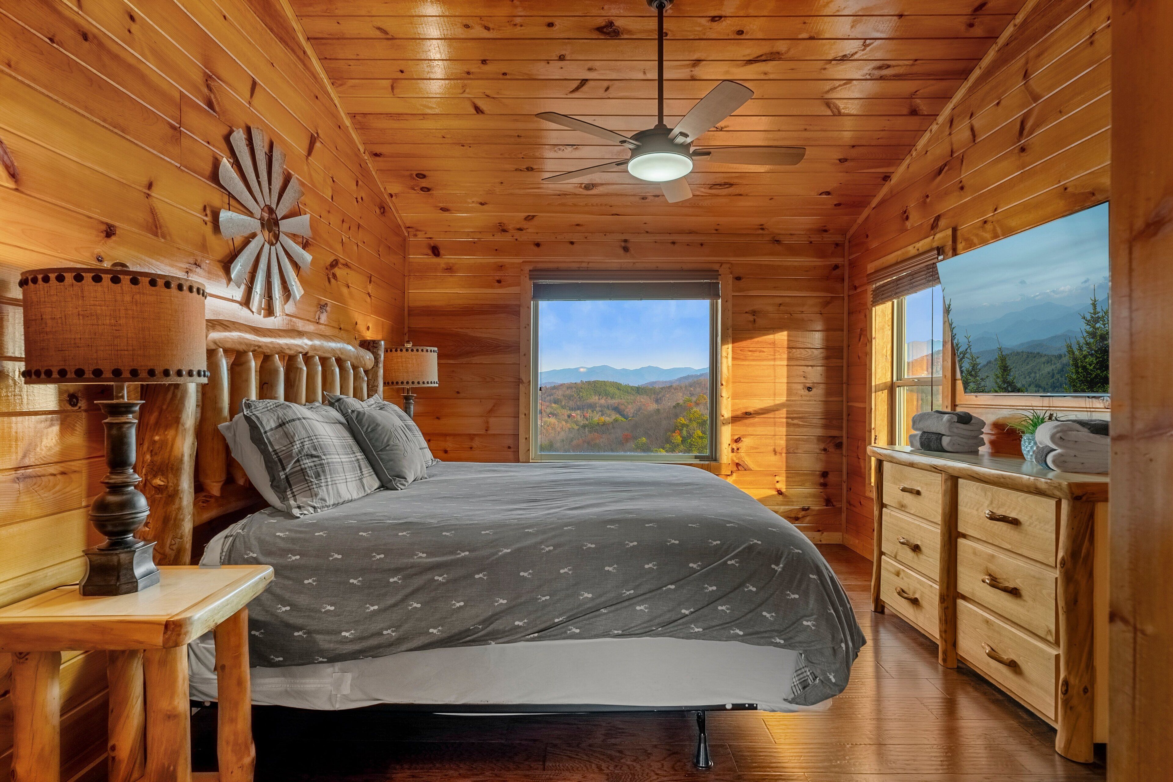Une chambre dans une cabane en bois, avec un lit, un ventilateur de plafond, une fenêtre donnant sur la montagne et une commode en bois.