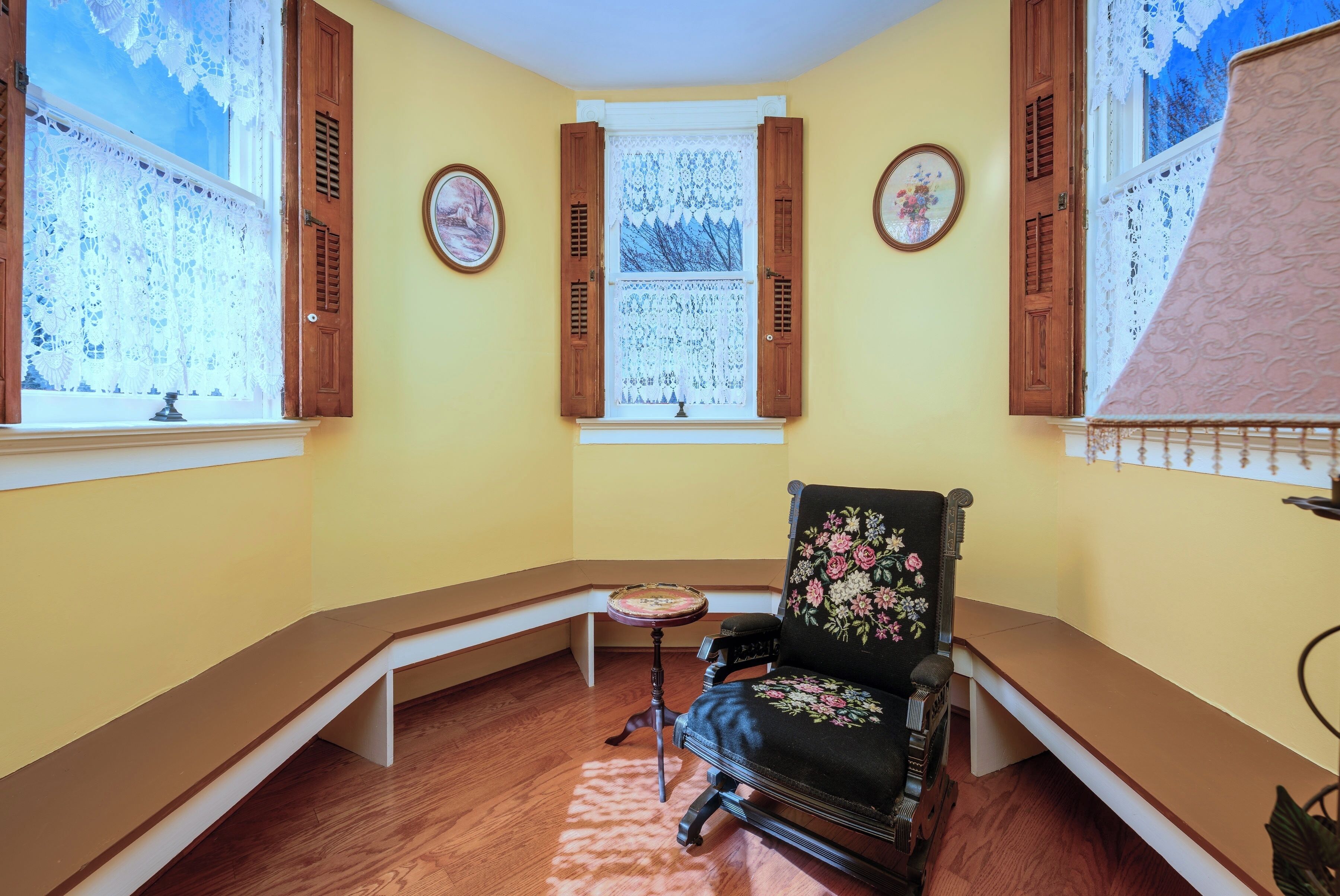 Una habitación en esquina con un banco de madera, un sillón floral, una mesita y una ventana con cortinas de encaje.