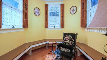 Una habitación en esquina con un banco de madera, un sillón floral, una mesita y una ventana con cortinas de encaje.