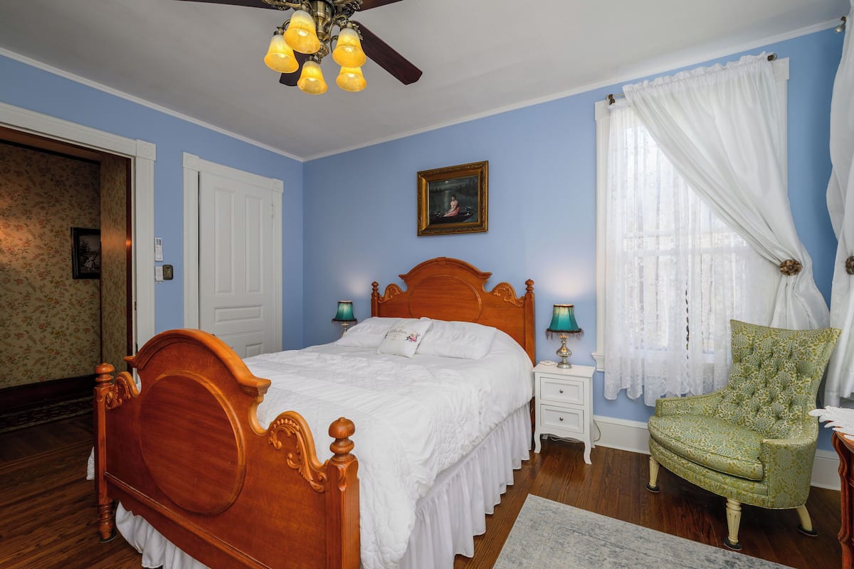 A bedroom with a wooden bed, a nightstand, a chair, a ceiling fan, and a window with curtains.