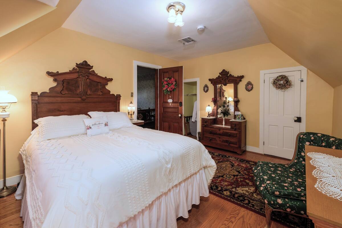 A bedroom with a large bed, wooden headboard, a dresser, a chair, and a door with a wreath.
