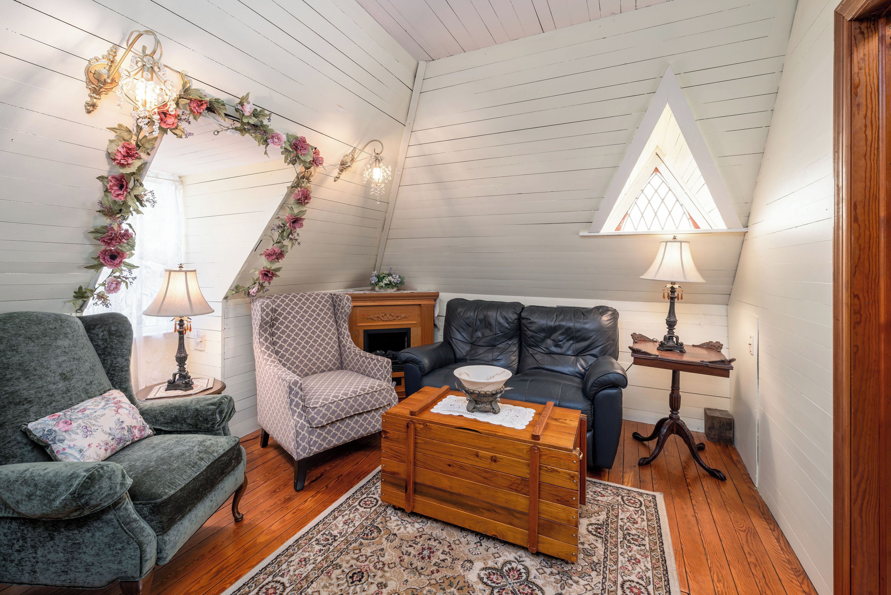 A cozy loft bedroom with a wooden ceiling, a fireplace, a blue leather sofa, a patterned armchair, and a wooden chest of drawers.