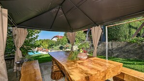 Mesa de madera al aire libre bajo una carpa, con vistas a una piscina y follaje.
