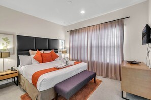 A modern hotel room with a large bed, a bedside table with a lamp, a bench, a TV, and a large window with curtains.