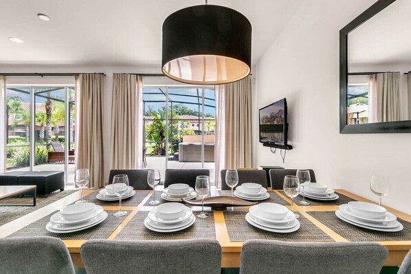 A hotel dining room with a large table set for a meal, a television on the wall, and large windows offering a view of the outdoors.