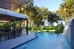 A swimming pool with a waterfall feature, surrounded by lush greenery and a modern building structure.
