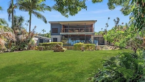 A modern two-story house with a large front lawn, surrounded by tropical vegetation.